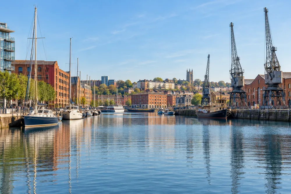 Boats moored on Bristol’s Floating Harbour beside brick waterfront buildings and dockside cranes
